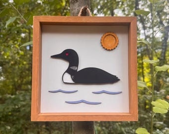May include: A square wooden frame containing a layered art piece featuring a black loon with a red eye swimming on light blue water waves. An orange sun is positioned in the upper right corner against a plain white background.