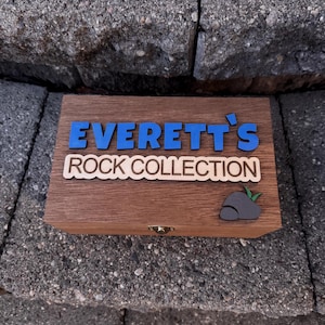 May include: A wooden box with the text "Everett's Rock Collection" in blue and white lettering. A small rock and leaf graphic is on the right side of the box. The box is sitting on a gray stone surface.