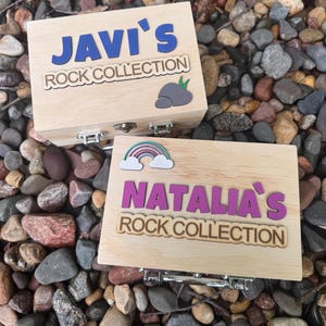 May include: Two rectangular light-colored wooden boxes with metal latches. One box features blue text reading JAVI'S ROCK COLLECTION with a gray rock icon. The other box features purple text reading NATALIA'S ROCK COLLECTION with a rainbow icon.