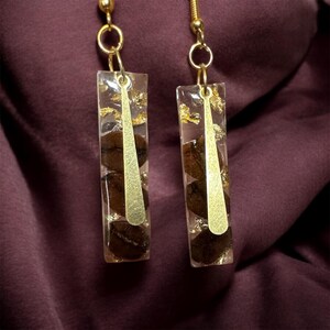 May include: A pair of rectangular dangle earrings. Each earring features coffee beans encased in clear resin, accented with gold leaf and a gold-colored metal accent. The earrings hang from gold-colored ear wires.