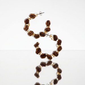 May include: A pair of hoop earrings with a unique design. Each earring is a clear resin hoop embedded with brown coffee beans and gold flakes. The earrings have silver-colored posts and are displayed on a reflective surface.
