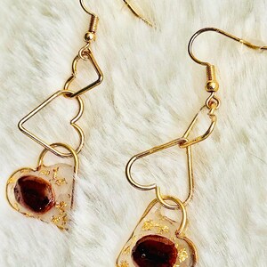 May include: Gold-colored dangle earrings. Each earring features a heart-shaped charm with a dark brown element and gold flakes encased in clear resin. The earrings have a heart and triangle design.