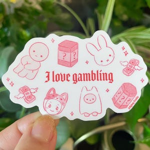 以下が含まれることがあります： ピンクと白のステッカーに「ギャンブルが好き」というテキストと、ウサギ、お金袋、サイコロ、帽子をかぶったキャラクターなど、さまざまなイラストが描かれています。