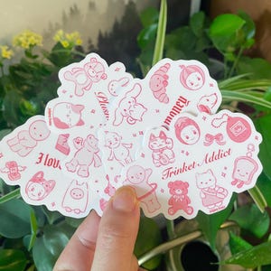 以下が含まれることがあります： ピンクと白のステッカーのセット。かわいいイラストとフレーズが描かれており、「大好き」「ぬいぐるみ」「お金」「小物中毒」「大好き」などがあります。猫、カエル、クマ、ウサギなど、さまざまなキャラクターが描かれています。