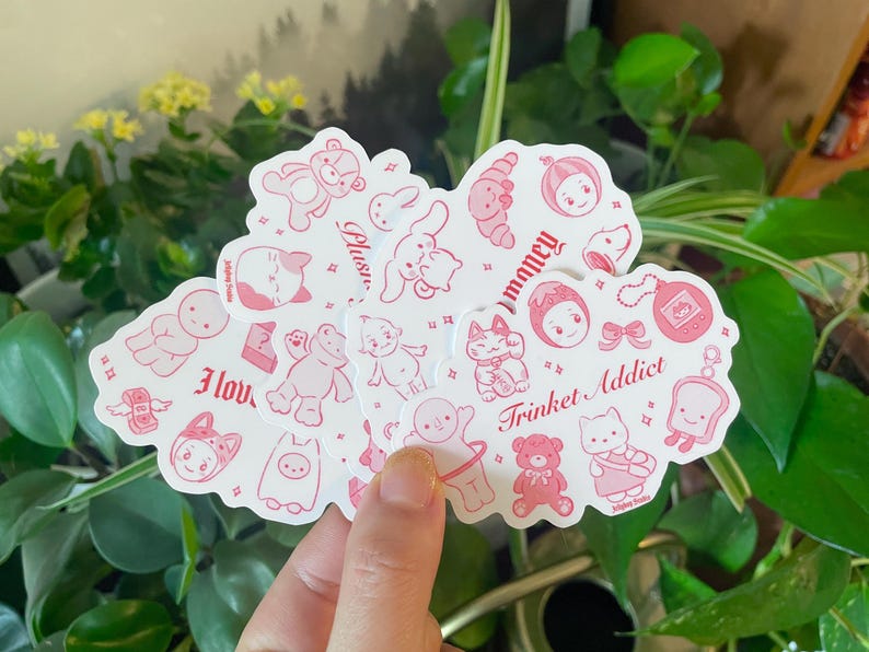 以下が含まれることがあります： ピンクと白のステッカーのセット。かわいいイラストとフレーズが描かれており、「大好き」「ぬいぐるみ」「お金」「小物中毒」「大好き」などがあります。猫、カエル、クマ、ウサギなど、さまざまなキャラクターが描かれています。