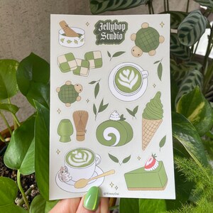 May include: A sheet of stickers featuring a green and white colour scheme with various designs including a matcha latte, a turtle, a whisk, a cake, and a matcha ice cream cone. The text "Jellybop Studio" is at the top of the sheet.