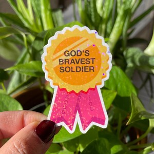 May include: A gold glitter sticker with a pink ribbon. The sticker says "God's Bravest Soldier".