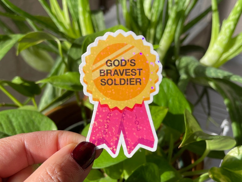 May include: A gold glitter sticker with a pink ribbon. The sticker says "God's Bravest Soldier".