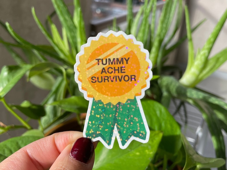 May include: A gold glitter sticker with a green glitter ribbon. The sticker says "Tummy Ache Survivor".