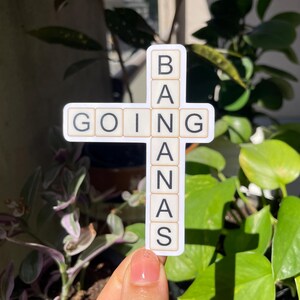 May include: A white sticker with the words "GOING BANANAS" spelled out in black letters on individual tiles. The tiles are arranged in a cross shape.
