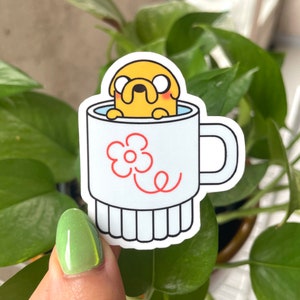 May include: A white ceramic mug with a red flower design and a yellow cartoon dog inside. The dog is wearing a blue collar and has a red nose.