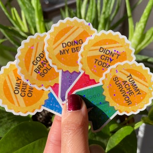 May include: A set of six yellow glitter stickers with motivational phrases. The phrases are "God's Brave Soldier", "Only Cried Once", "Doing My Best", "Didn't Cry Today", "Tummy Ache Survivor", and "Good Job".