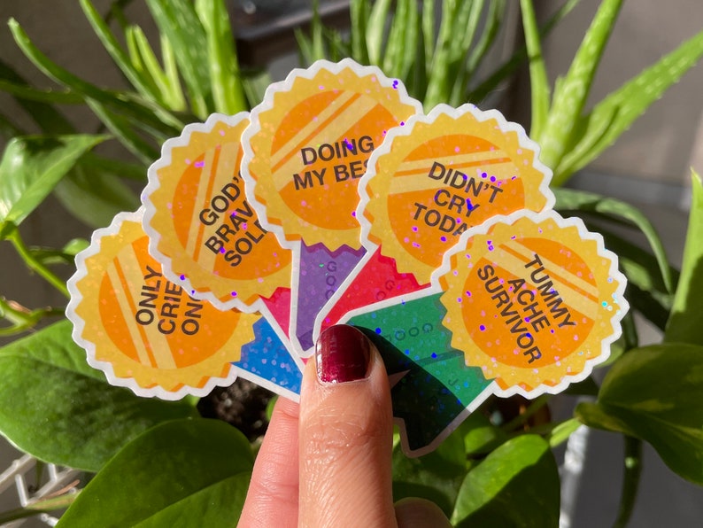 May include: A set of six yellow glitter stickers with motivational phrases. The phrases are "God's Brave Soldier", "Only Cried Once", "Doing My Best", "Didn't Cry Today", "Tummy Ache Survivor", and "Good Job".