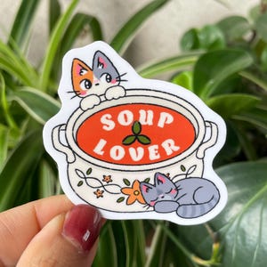 May include: A white sticker featuring a cartoon soup pot with the words "SOUP LOVER" in orange. Two cats are depicted, one peeking over the pot and another sleeping below. The soup is orange, and the pot has floral designs.