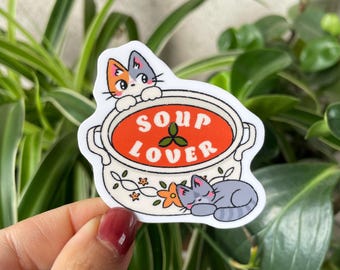 May include: A white sticker with a cartoon soup pot design. The pot contains orange soup and the words "SOUP LOVER". Two cats are featured, one looking over the pot and another sleeping. The pot has floral designs.