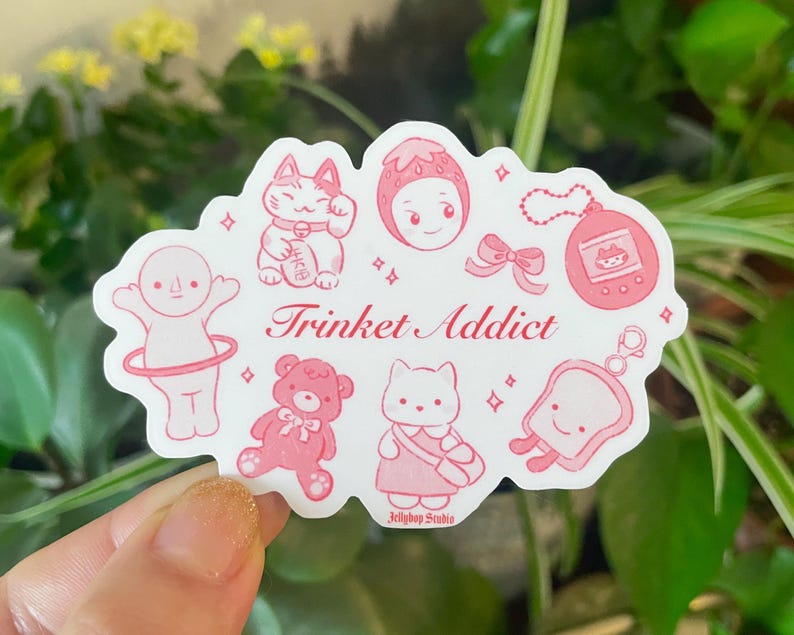 以下が含まれることがあります： ピンクと白のステッカーに「Trinket Addict」の文字と、猫、イチゴ、フープを持った人、テディベア、バッグを持った猫、パンの切れ端など、さまざまなかわいいイラストが描かれています。