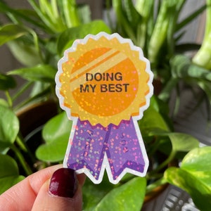 May include: A gold glitter sticker with a purple ribbon. The sticker says "Doing My Best".
