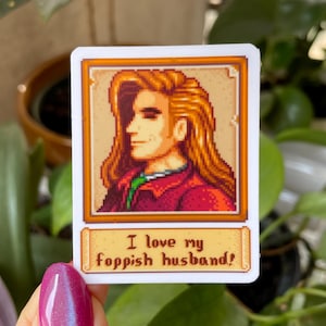 May include: A sticker featuring a pixelated portrait of a man with long blonde hair. The portrait is framed in gold and has a scroll at the bottom that reads "I love my foppish husband!"