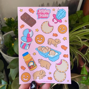 May include: A pink sheet of paper with various stickers on it. The stickers are of different shapes and sizes and feature various images, including a chocolate bar, a carton of ice cream, a ring with a blue gem, a lion, an elephant, and a juice box. The text "Jellybop Studio" is printed in black at the top of the sheet.