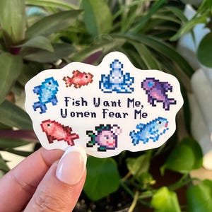 May include: A white sticker featuring pixelated fish illustrations in blue, red, and purple. The text "Fish Want Me, Women Fear Me" is printed in black. The sticker is surrounded by green foliage.