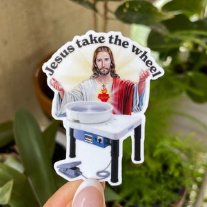 May include: A sticker featuring an image of Jesus with the text "Jesus take the wheel." Jesus is depicted with a glowing halo, red and white robes, and a sacred heart. Below him is a pottery wheel on a white table.
