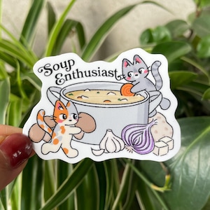 May include: A sticker featuring a cartoon illustration of a soup pot with the text "Soup Enthusiast". Two cats are near the pot, and vegetables like carrots, onions, and garlic are also shown. The sticker is white with a glossy finish.