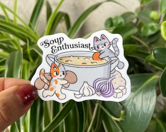 May include: A sticker with a cartoon illustration of a soup pot and the text "Soup Enthusiast". Two cats are depicted near the pot, alongside vegetables such as carrots, onions, and garlic. The sticker is white with a glossy finish.