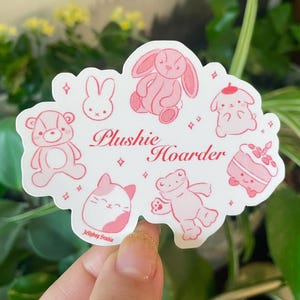 以下が含まれることがあります： ピンクと白のステッカーに「Plushies Hoarder」の文字と、ウサギ、カエル、猫、クマ、ケーキ、鳥など、さまざまなぬいぐるみが描かれています。ステッカーは、かわいらしく幻想的なスタイルでデザインされています。
