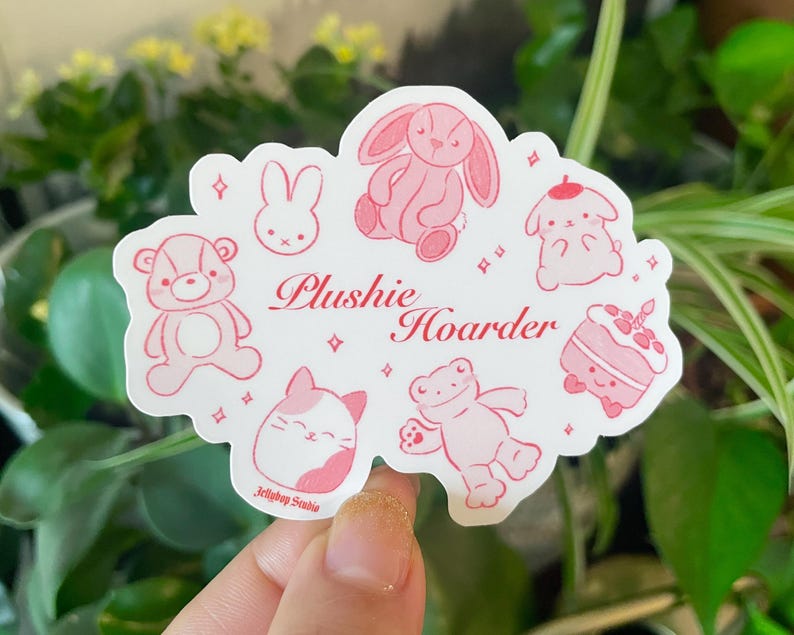 以下が含まれることがあります： ピンクと白のステッカーに「Plushies Hoarder」の文字と、ウサギ、カエル、猫、クマ、ケーキ、鳥など、さまざまなぬいぐるみが描かれています。ステッカーは、かわいらしく幻想的なスタイルでデザインされています。