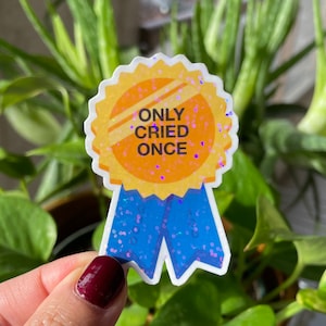 May include: A gold glitter sticker with a blue glitter ribbon. The sticker says "ONLY CRIED ONCE".