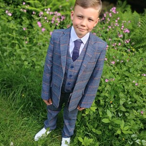 May include: A young person wearing a blue plaid suit with a light blue shirt and a dark blue tie. The person is standing in a grassy area with pink flowers.