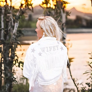 May include: A white denim jacket with fringe detailing and silver stars. The jacket has the word "WIFEY" written on the back.