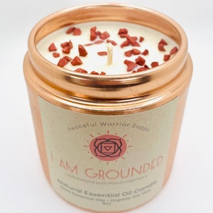May include: A copper candle jar with a white candle and red crystals. The label reads "Peaceful Warrior Soaps" and "I AM GROUNDED Cedarwood.patchouli.rosemary Natural Essential Oil Candle 100% Essential Oils. Organic Soy Wax 8oz."