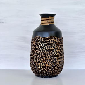 May include: A black and gold decorative vase. The vase has a textured, patterned lower half with a gold interior. The top portion is black with a gold band around the neck. The vase sits on a white surface.