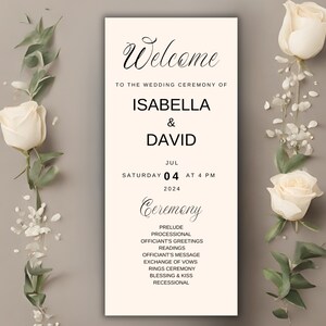 May include: A wedding ceremony sign with the names Isabella & David, date, and time. The sign is cream-colored with black text and floral accents. White roses and greenery frame the sign.
