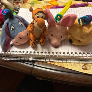 Könnte beinhalten: Ein Set aus vier Plüsch-Schlüsselanhängern mit I-Aah, Tigger, Ferkel und Winnie Puuh. Jedes Plüschtier hat einen bunten Kunststoffclip an der Oberseite und zeigt die ikonischen Gesichter und Formen dieser bekannten Figuren aus Kindergeschichten.