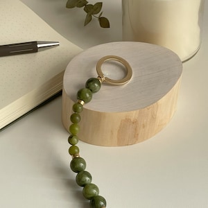 May include: A green jade beaded keychain with a gold ring and a gold connector. The keychain is displayed on a wooden surface.
