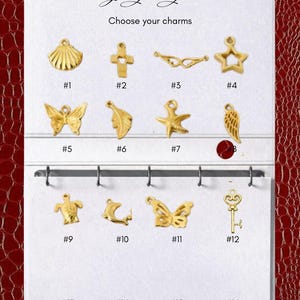 May include: A white display card with the text Design your journal and Choose your charms, featuring twelve gold-toned metal charms numbered 1 through 12. The charms include shapes like a shell, cross, wings, star, butterfly, leaf, starfish, feather, turtle, dolphin, and key.