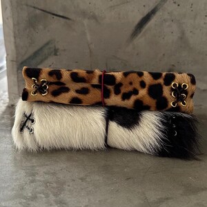 May include: Two rectangular cuffs made of animal hair material. The top cuff features a brown and black leopard print pattern with gold-toned eyelets and dark brown lacing. The bottom cuff is white with black patches and silver-toned eyelets.