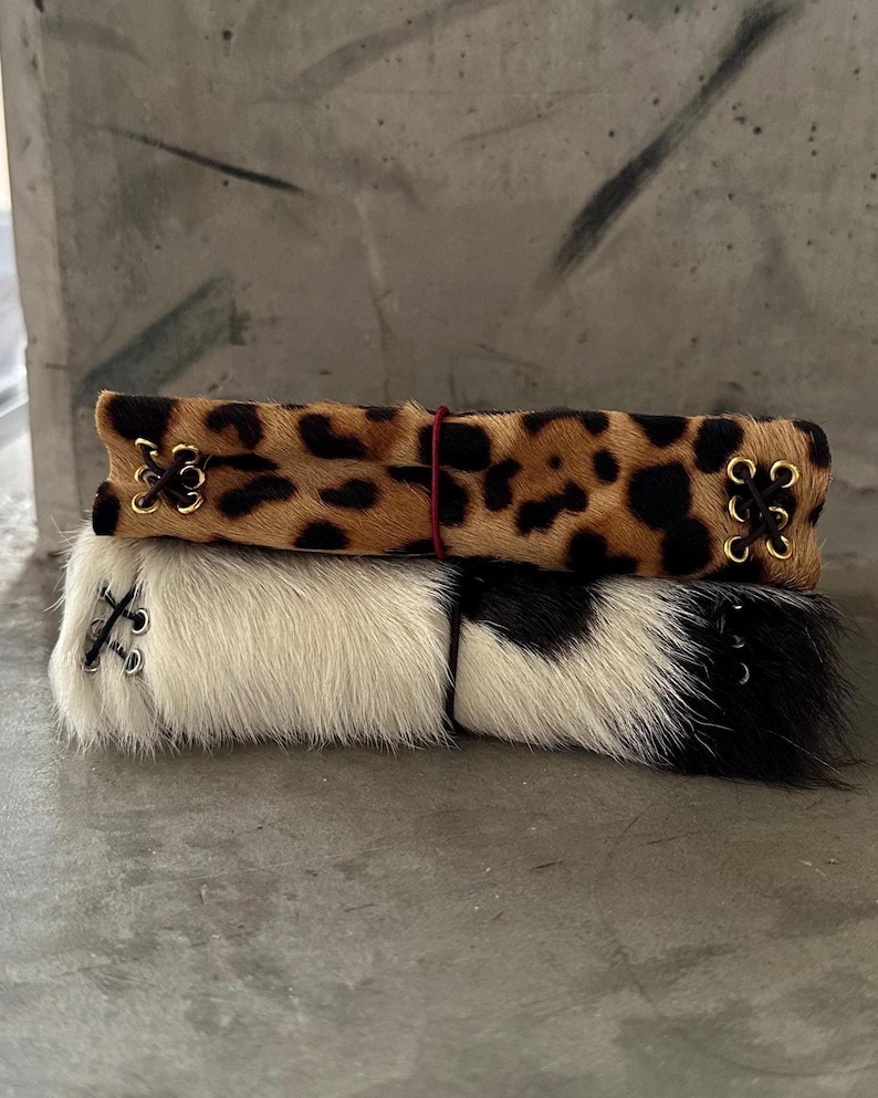 May include: Two rectangular cuffs made of animal hair material. The top cuff features a brown and black leopard print pattern with gold-toned eyelets and dark brown lacing. The bottom cuff is white with black patches and silver-toned eyelets.