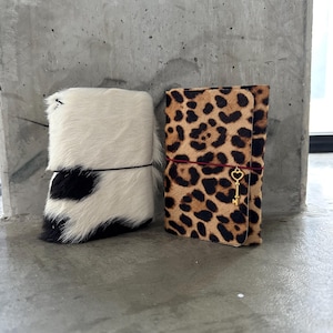 May include: Two rectangular notebooks with textured hair-on-hide covers. The left notebook features a black and white cow print pattern with a thin black elastic band. The right notebook has a brown and black leopard print with a red elastic band and a gold-toned key charm.