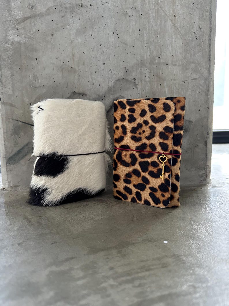 May include: Two rectangular notebooks with textured hair-on-hide covers. The left notebook features a black and white cow print pattern with a thin black elastic band. The right notebook has a brown and black leopard print with a red elastic band and a gold-toned key charm.
