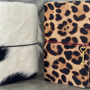 May include: Two rectangular journals with textured hair-on-hide covers. One journal features a black and white cow print with a thin black elastic band. The other journal has a tan and black leopard print with a red elastic band and a gold key charm.