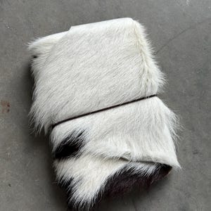 May include: A rectangular journal or notebook covered in white and black cowhide fur. The cover features a long-haired texture with a distinct white section on top and a dark black patch at the bottom, secured by a thin, dark brown elastic band.