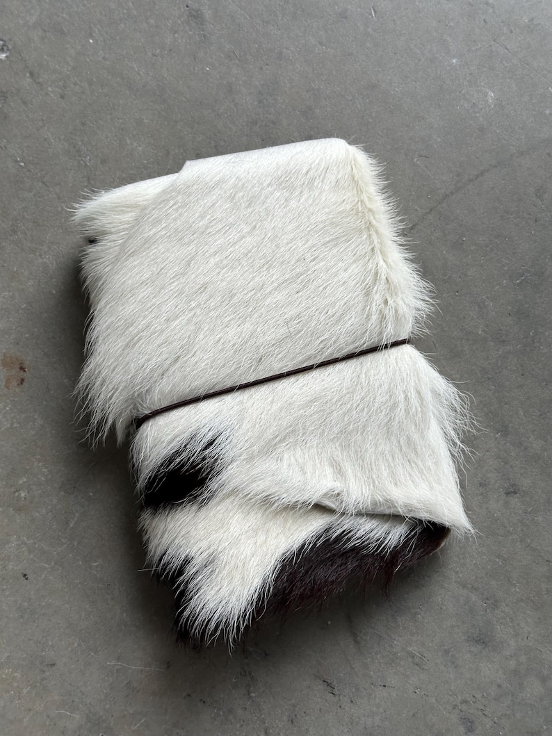 May include: A rectangular journal or notebook covered in white and black cowhide fur. The cover features a long-haired texture with a distinct white section on top and a dark black patch at the bottom, secured by a thin, dark brown elastic band.