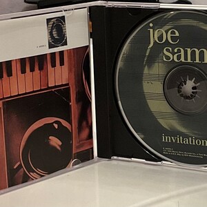 May include: A CD case with a black and white image of a piano keyboard and a black and white image of a record player on the front cover. The CD case is open and the CD is visible. The CD is a light gray color with a black spiral in the center. The CD has the text 'joe sample' in yellow letters and the text 'invitation' in white letters.