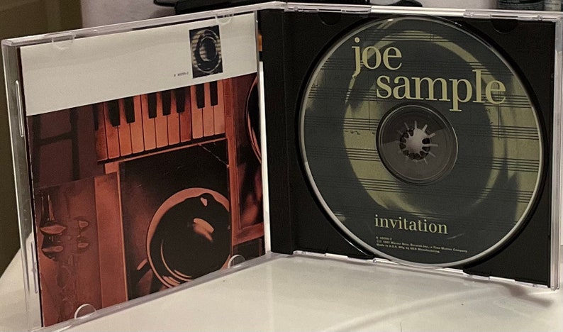 May include: A CD case with a black and white image of a piano keyboard and a black and white image of a record player on the front cover. The CD case is open and the CD is visible. The CD is a light gray color with a black spiral in the center. The CD has the text 'joe sample' in yellow letters and the text 'invitation' in white letters.