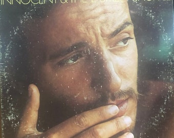 May include: A vintage vinyl record cover featuring a close-up of Bruce Springsteen's face. The cover is black and white with the title "Bruce Springsteen: The Wild, The Innocent & The E Street Shuffle" printed in white.