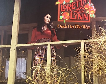 Pode incluir: Uma capa de disco de vinil vintage com a cantora de música country Loretta Lynn. A capa mostra Lynn em pé em uma varanda, olhando para o espectador. O título do álbum, "One's On The Way", está impresso em letras grandes abaixo dela.