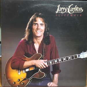 May include: Album cover featuring Larry Carlton holding a sunburst electric guitar. He wears a burgundy shirt and smiles at the viewer. The album title "Sleepwalk" is in white and red text.